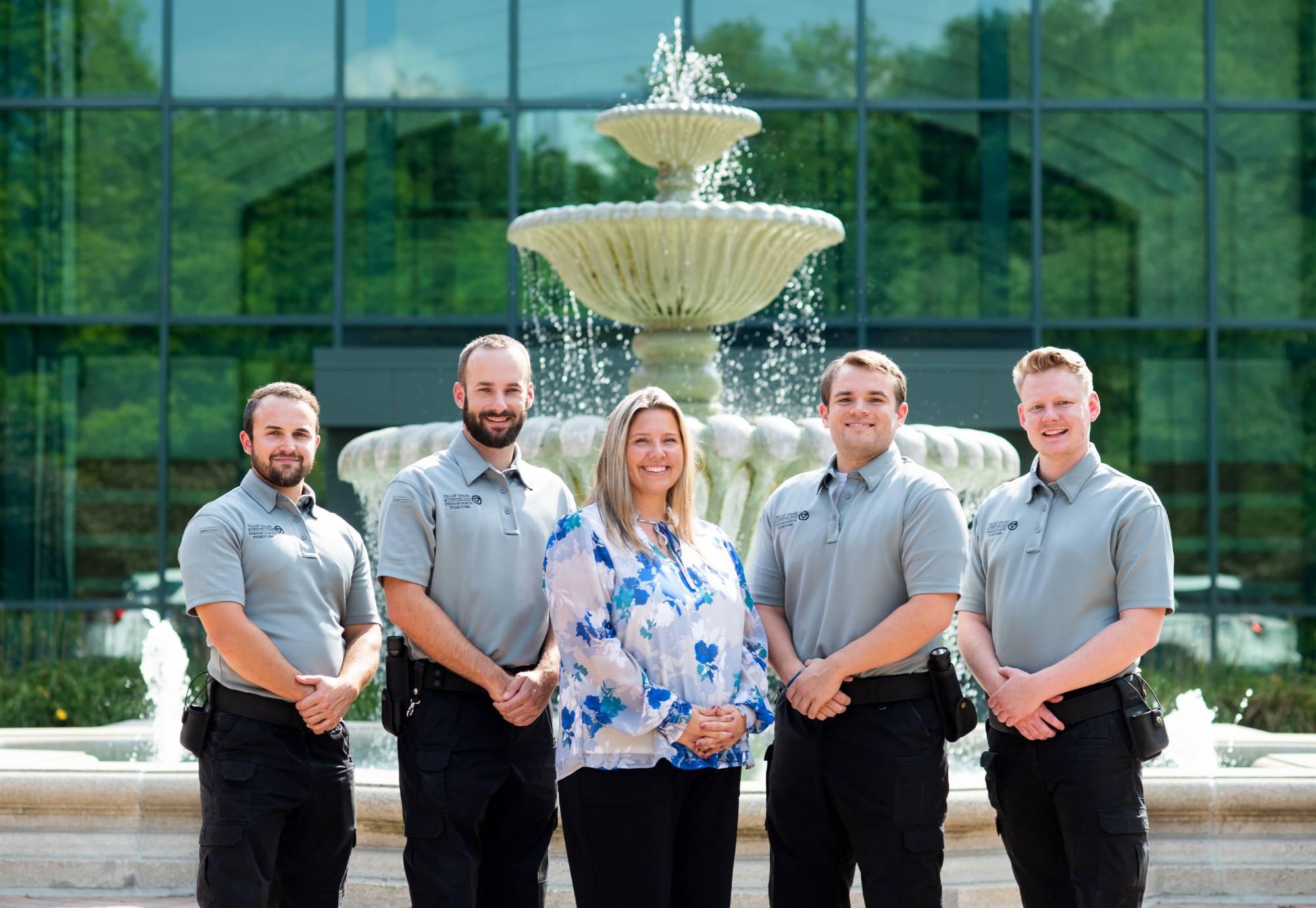 Security employees, 4 in uniform, and 1 supervisor in plain clothes, posing in front of the Student Services fountain. Subjects are smiling.